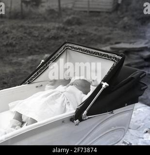 1958, historique, à l'extérieur dans un landau ou une babycalèche de l'époque, un nouveau-né couché sur le dos dormant. Pour une respiration optimale et pour obtenir tout l'oxygène dont ils ont besoin, les nouveaux bébés doivent être à plat et le pram classique construit en autocar avec sa grande capuche, son cadre généreux et son intérieur entièrement plat leur permettent de faire cela avec beaucoup d'espace. Cette position allongée a également encouragé leur colonne vertébrale et leurs hanches à se développer correctement. Banque D'Images