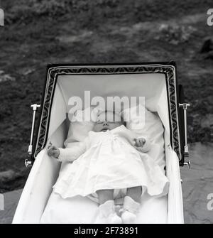 1958, historique, à l'extérieur dans un landau ou un babycalèche construit en autocar de l'époque, un nouveau-né allongé sur son dos dormant paisiblement. Pour une respiration optimale et pour obtenir tout l'oxygène dont ils ont besoin, les nouveaux bébés doivent être à plat et le pram classique construit en autocar de cette époque, avec sa large capuche, son cadre généreux et son intérieur entièrement plat, ils peuvent le faire avec beaucoup d'espace. Cette position allongée favorise également le développement correct de la colonne vertébrale et des hanches. Banque D'Images