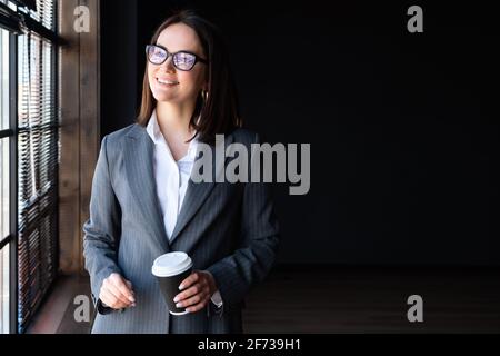 Jeune femme en costume d'affaires avec une boisson dans un verre de papier se tient près de la fenêtre. Banque D'Images
