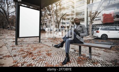 Un homme à la mode et à la fois élégant et élégant le costume et les lunettes sont assis sur le banc à l'intérieur de un arrêt de bus de ville avec un blanc adv Banque D'Images