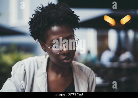 Un portrait d'une jeune femme noire aux cheveux bouclés dans un café de rue ; une charmante femme africaine présente des spectacles et une tranchée blanche lit un menu Banque D'Images