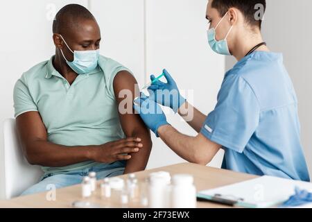 Jeune médecin dans le masque de protection faire l'injection à l'âge moyen patient afro-américain Banque D'Images