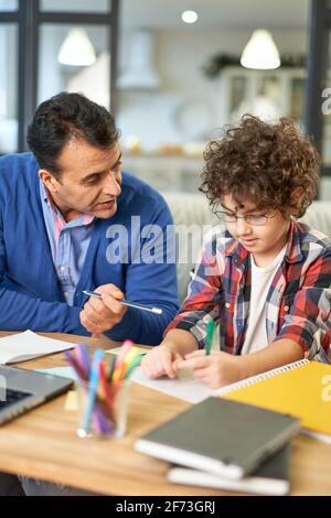 Meilleur enseignant. Un père hispanique affectueux aidant son fils à faire ses devoirs, expliquant la tâche tout en étant assis à son bureau à la maison Banque D'Images