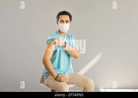 Un homme avec un masque médical et un bracelet sur son épaule est assis sur une chaise sur fond gris. Banque D'Images