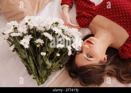 Une belle brunette dans une robe rouge se trouve sur le lit à côté d'un bouquet de pâquerettes Banque D'Images