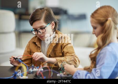 Portrait d'un garçon mignon portant des lunettes et expérimentant avec l'électricité des circuits tout en construisant des robots pendant les cours d'ingénierie à l'école Banque D'Images