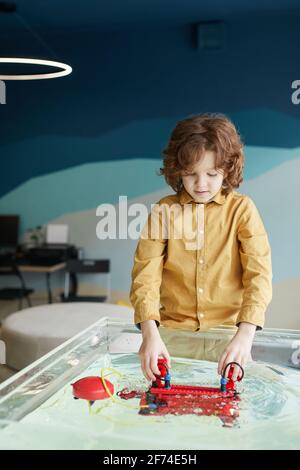 Portrait vertical d'un joli petit garçon aux cheveux bouclés mettant le bateau à rabo dans l'eau tout en expérimentant la technologie au laboratoire de robotique dans école Banque D'Images