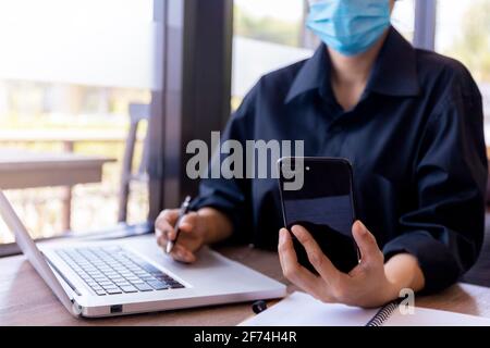 Jeune femme avec masque de protection assis dans un café à table en bois à l'aide d'un smartphone.et d'un ordinateur portable. Fille de navigation sur Internet, de chat, de blogging. FE Banque D'Images