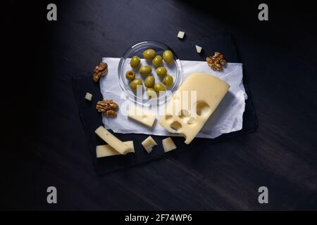 fromage semi-dur avec trous, maasdam coupé en tranches se trouve sur une assiette de pierre avec des olives et des noix sur fond sombre Banque D'Images