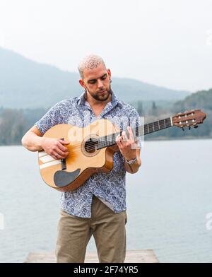 Homme barbu dans des vêtements décontractés debout avec guitare sur bois jetée près de la rivière avec des montagnes sur fond gris nuageux ciel en journée Banque D'Images