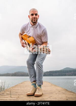 Corps complet de barbu gars dans des vêtements décontractés debout avec guitare sur une jetée en bois près de l'herbe et de la rivière avec des montagnes sur fond ciel gris nuageux Banque D'Images