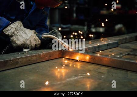 Travailleur sans visage en gants et en métal de soudage uniforme sur la table près des constructions en usine Banque D'Images