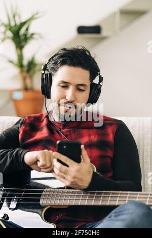 Musicien masculin barbu adulte avec guitare basse dans le texte du casque messagerie sur téléphone portable dans la salle de séjour Banque D'Images