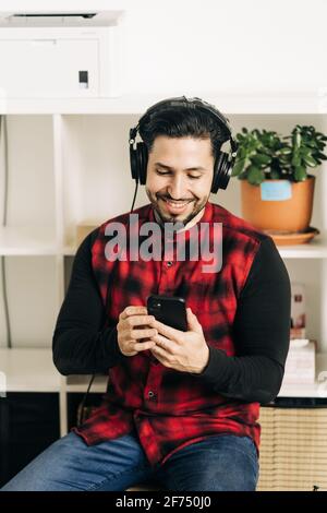 Musicien masculin barbu adulte dans un casque de messagerie texte sur un téléphone portable dans la salle de séjour Banque D'Images