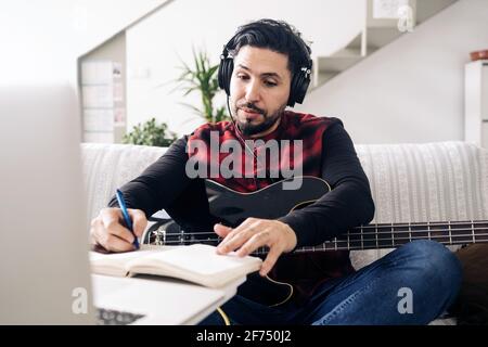 Guitariste mâle adulte dans un casque avec guitare électrique prenant des notes dans l'ordinateur portable tout en composant de la musique contre netbook sur le canapé à accueil Banque D'Images