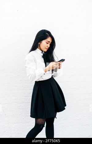 Vue latérale de la jeune femme ethnique dans une tenue habillée debout près du mur blanc et à l'aide d'un téléphone portable Banque D'Images
