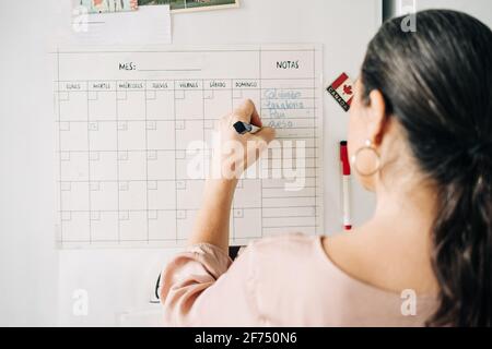 Vue arrière de la femme anonyme prenant des notes dans le calendrier sur réfrigérateur avec aimants tout en faisant des plans dans la cuisine à la maison Banque D'Images