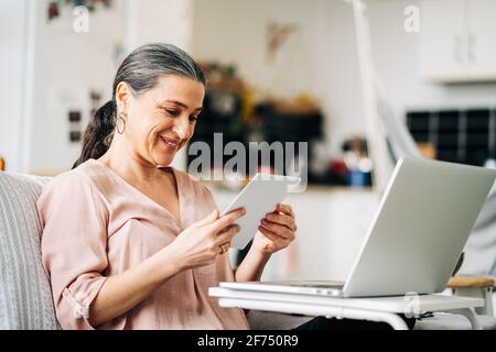 Femme d'âge moyen et gaie assise sur un canapé près d'un netbook moderne tout en regardant la photo dans un appartement moderne avec cuisine arrière-plan flou Banque D'Images