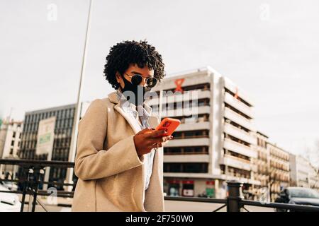 De dessous de la femme afro-américaine élégante dans le masque en tissu envoi de sms sur un téléphone portable en marchant dans la rue ville Banque D'Images