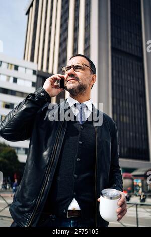 Homme hispanique mature Executive dans une tenue élégante et décontractée avec chaud buvez pour parler sur votre téléphone portable tout en vous promenant en ville chaussée Banque D'Images
