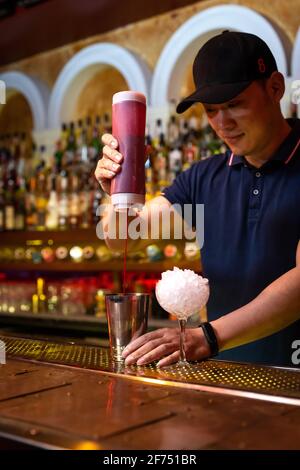 Jeune barman asiatique versant du jus de fraise dans le shaker pendant préparation d'un cocktail au bar Banque D'Images