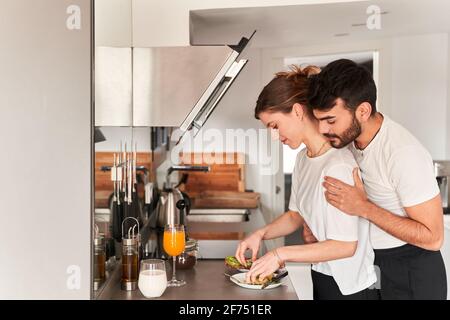 Vue latérale d'un jeune homme ethnique romantique en blanc chemise souriante et enserrer bonne petite amie de préparer un petit déjeuner sain avec avocat dans la cuisine Banque D'Images