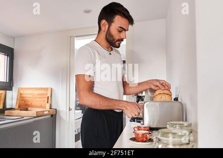 Vue latérale d'un jeune homme hispanique barbu dans des vêtements décontractés faire des toasts croquants tout en préparant le petit déjeuner à la maison le matin Banque D'Images