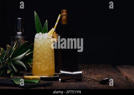 Verre de cocktail alcoolisé décoré de morceaux d'ananas et de feuilles avec de la paille sur le bac près de l'agitateur et bouteille avec tirer le verre à la table sur le bac Banque D'Images