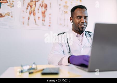 Jeune Afro-américain concentré médecin masculin dans la robe médicale et Les écouteurs TWS fonctionnent sur un ordinateur portable tout en étant assis à la table clinique moderne Banque D'Images