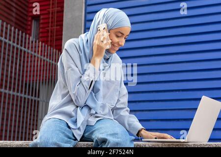 Femme musulmane souriante en foulard avec netbook parlant sur téléphone portable en ville Banque D'Images