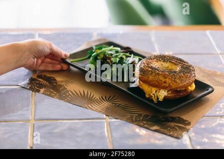 Grand angle de récolte anonyme personne servant délicieux sandwich bagel avec du fromage et du poulet sur l'assiette à table dans le café Banque D'Images