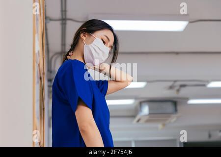 Jeune femme asiatique fatiguée et frustrée portant l'uniforme et masque chirurgical souffrant de douleurs au cou pendant le travail à l'hôpital au cours de l'épidémie de covid-19 Banque D'Images