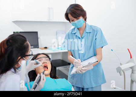 Dentiste examinant les dents de la patiente lorsque l'infirmière prend des notes dans le document Banque D'Images