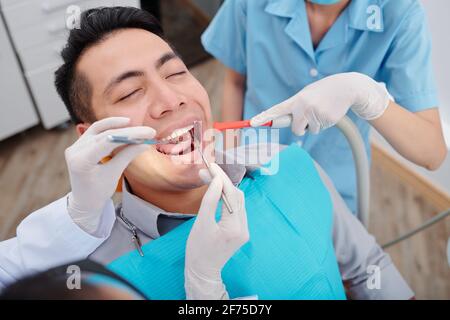 Dentiste traitant les dents d'un patient mâle lorsque l'infirmière met de la salive éjecteur à l'intérieur de la bouche Banque D'Images