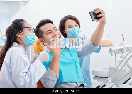 Jeune homme asiatique heureux avec son nouveau sourire prenant selfie avec dentiste et son assistant pour les médias sociaux Banque D'Images