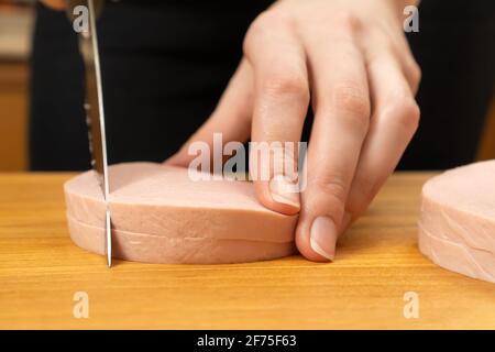 Les mains des femmes ont coupé une saucisse bouillie en cubes sur une planche de bois avec un couteau de gros plan. Banque D'Images