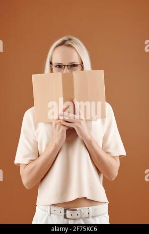 Souriant timide jolie femme étudiante à l'université couvrant la face avec ouvert réservez et regardez l'appareil photo Banque D'Images