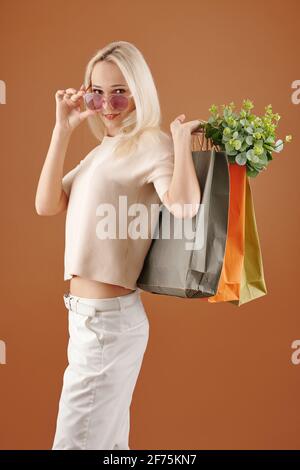 Bonne jeune femme positive avec des sacs de shopping en papier qui se départir lunettes de soleil et regarder l'appareil photo Banque D'Images