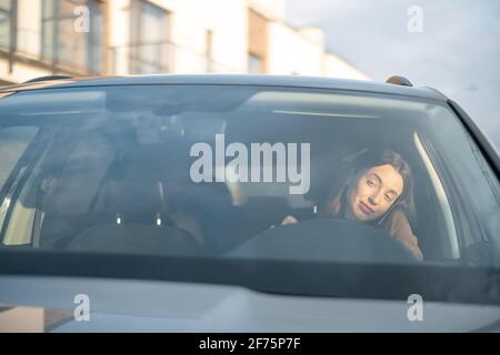 Une jeune femme parle au téléphone tout en conduisant une voiture et en tenant un téléphone sur son épaule. Femme d'affaires qui va travailler de la maison. Vue par le pare-brise. Banque D'Images