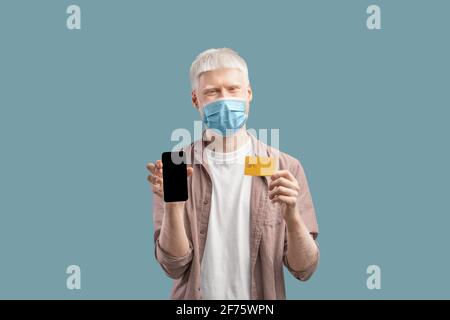 Albino Guy dans le masque de protection tenant le smartphone et la carte de crédit sur fond de studio turquoise Banque D'Images