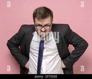 Homme caucasien barbu dans des lunettes et un costume d'affaires est embûté et regarde furtivement dans la caméra. Fond rose. Banque D'Images