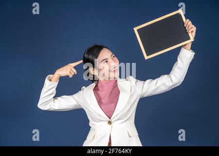 Jeune femme asiatique souriante tenant un espace de copie vide message tableau noir sur fond bleu, pensant et taquisant via ballon papier blanc, P Banque D'Images