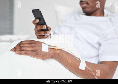 Gros plan de l'homme afro-américain dans le lit d'hôpital et en utilisant un smartphone avec l'accent sur le cathéter de perfusion iv dans la main, copier l'espace Banque D'Images