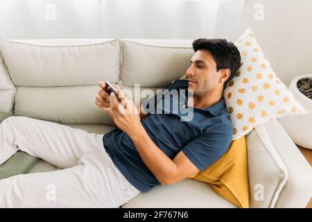Beau jeune homme caucasien allongé sur un canapé dans un confortable permet de jouer à des jeux et de regarder des vidéos sur votre smartphone pendant la période libre temps à la maison Banque D'Images