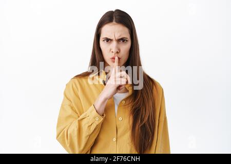 Shush, Taisez-vous. Une fille bossy sérieuse qui se précipite, dit de garder la bouche fermée, geste tabou, demandant le silence, se comporter shhh, se tenant sur blanc Banque D'Images