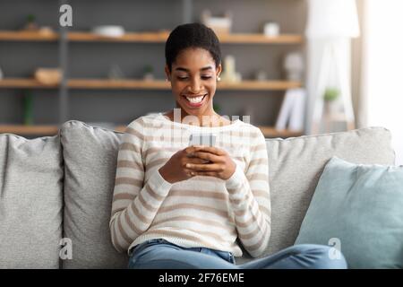 Joyeuse Black Lady détente avec un smartphone dans le salon, messagerie avec des amis Banque D'Images