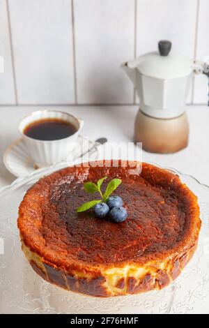 Gâteau au fromage basque maison brûlé sur une assiette avec myrtilles et feuilles de menthe sur fond clair, avec une tasse de café et une cafetière geyser. Banque D'Images