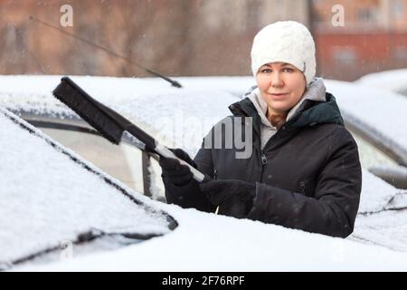 Une femme utilisant une brosse pour enlever la neige et la glace du pare-brise et du capot de sa voiture, l'hiver Banque D'Images