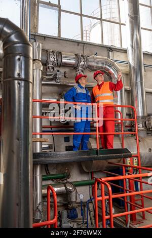 L'ingénieur de maintenance se tient à côté de l'ouvrier du pétrole dans la raffinerie de pétrole avec de grandes canalisations. Banque D'Images
