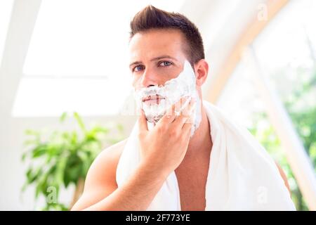 Prise de vue d'un beau homme avec une serviette autour de son cou à l'aide de mousse de rasage avant de se raser en se tenant devant le miroir de la salle de bains. Banque D'Images